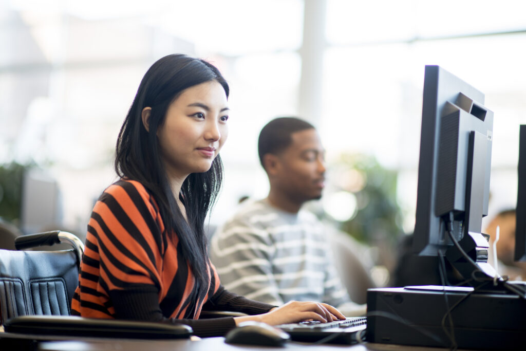 Woman in forefront of shot, working on computer designing video game. Co-worker can be seen in background, also working on a game.